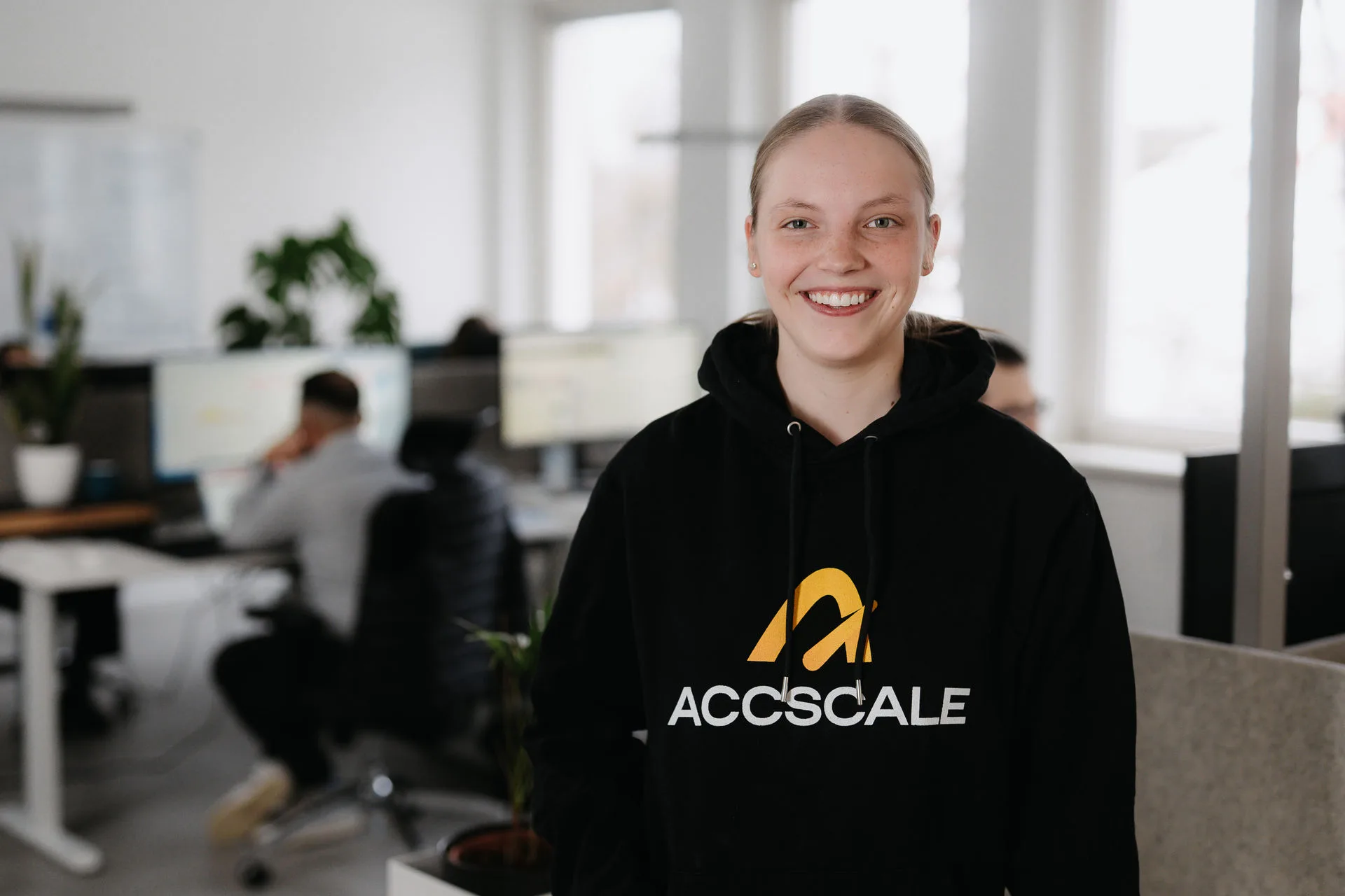 Kollegin im ACCSCALE-Hoodie lächelt im Büro, im Hintergrund Arbeitsplätze